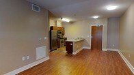 This single-floor unit offers an open and functional layout that seamlessly connects the kitchen and living areas. The kitchen is equipped with stainless steel appliances, including a refrigerator, microwave, and range, complemented by rich wood cabinetry and granite-style countertops. A peninsula breakfast bar extends from the kitchen, providing additional prep space and casual seating while maintaining an open sightline to the living area. Warm hardwood flooring spans the entirety of the space, lending a cohesive and inviting feel throughout. The living area is generously sized, offering flexible furniture arrangement possibilities. Neutral-toned walls provide a clean backdrop that suits a variety of décor styles. The unit features a dedicated entryway with a solid wood door, offering a clear sense of arrival. Climate control is handled via in-wall vents, and the unit is equipped with a thermostat for personalized comfort. Overhead lighting provides consistent illumination throughout. The overall layout is efficient and practical, making excellent use of the available space while maintaining an open and airy ambiance ideal for everyday living.