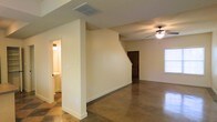 This two-story property offers a thoughtfully designed layout with distinct character throughout. The main level showcases polished concrete floors that lend an industrial-chic aesthetic to the open living area, which flows seamlessly into the surrounding spaces. A ceiling fan overhead provides both function and a grounding focal point to the room, while a window allows natural light to filter in and brighten the interior. Neutral, cream-toned walls throughout create a clean and versatile backdrop, complementing the warm concrete underfoot. A staircase with a dark wood door nearby connects the main level to the upper floor, adding a sense of vertical dimension to the home. Visible HVAC vents in the ceiling indicate a central climate control system integrated into the architecture. Adjacent to the main living space, a hallway leads to an additional room—visible in the background—featuring tiled flooring, suggesting a bathroom or utility space. The overall ambiance is open, practical, and move-in ready, with a neutral palette that allows for a wide range of interior design possibilities across both floors.