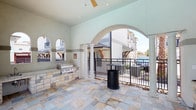 This covered outdoor lounge offers a welcoming space designed for socializing and leisure. The area is defined by a series of graceful arched openings supported by sturdy columns, framing views of the surrounding community and creating an open, airy atmosphere. The flooring consists of natural stone-style tiles laid in a multicolored pattern, adding a refined, durable surface underfoot.

A built-in grill station anchored along one wall features a stone surround, making it a functional centerpiece for outdoor cooking and entertaining. A ceiling fan overhead ensures comfortable airflow throughout the covered space, enhancing usability across varying weather conditions. A high-top bistro table provides a casual spot for conversation or dining, complemented by a nearby waste receptacle for convenience.

Black iron railing lines the perimeter, offering a defined boundary while maintaining open sightlines to adjacent outdoor areas, including a pool visible in the background. The soft green and neutral tones of the walls contribute to a calm, inviting ambiance. Overall, the lounge combines practical amenities with an attractive design, creating an ideal setting for outdoor gatherings.