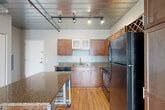 Located at 201 S 1st St in Ann Arbor, MI, this one-bedroom, one-bathroom loft offers a distinctive industrial aesthetic defined by exposed metal ductwork and a corrugated metal ceiling, creating a modern urban character throughout. Rich hardwood flooring runs through the main living areas, while track lighting adds a stylish, functional touch.

The open-concept kitchen is a standout feature, showcasing warm wood cabinetry, granite countertops, black appliances including a range, microwave, and refrigerator, and a built-in wine rack. A mobile granite-topped kitchen island provides additional prep space and flexibility. The kitchen flows seamlessly into the living area, which is bathed in natural light from large windows offering views of the surrounding Ann Arbor streetscape.

The bedroom is carpeted for comfort and features generous windows that bring in ample daylight and outdoor views. It connects to a dedicated closet with hanging storage. Two additional closets and a utility room provide practical storage throughout the unit.

The bathroom is finished with a wood vanity, a white vessel sink, and a soaking tub with a shower combination. In-unit laundry adds further convenience. The entrance hallway completes this well-organized, efficiently designed urban loft.