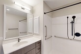 Located at 405 Lake Lida Lane in Rochelle, Illinois, this efficiently designed one-bedroom, one-bathroom unit offers a clean, modern aesthetic across a single floor. The layout features a hallway connecting the bedroom and bathroom, finished with durable grey luxury vinyl plank flooring and crisp white panel doors with sleek brushed-nickel hardware throughout.

The bedroom is a well-proportioned private retreat with neutral-toned walls, a window providing natural light, and ample space for essential furnishings. Its understated design keeps the focus on functionality and comfort.

The bathroom is a standout feature of this unit, thoughtfully designed with accessibility in mind. It includes a large roll-in shower equipped with a handheld showerhead, matte black fixtures, and multiple stainless steel grab bars for added safety. A spacious vanity with a white quartz countertop, an undermount sink, and wood-toned cabinetry with black hardware provides practical storage. A large mirror with overhead bar lighting brightens the space, while a recessed medicine cabinet adds additional storage. The toilet area features a wall-mounted grab bar, and the floor is finished with light tile for a clean, polished look.

Overall, the unit presents a contemporary, accessible design with a neutral color palette, modern fixtures, and thoughtful details that prioritize both style and practicality.