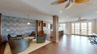 Located on Village Green Blvd in Aiken, South Carolina, this thoughtfully designed single-floor space offers a cohesive blend of modern aesthetics and warm, inviting interiors. Rich hardwood flooring flows throughout, anchoring the open layout and connecting each distinct area seamlessly.

The main lounge area is a visual centerpiece, highlighted by a bold blue-and-white chevron accent wall and warm vertical wood slat paneling. Teal leather seating arranged around a round coffee table creates an intimate conversation setting, while a sculptural pendant light adds a refined finishing touch. Adjacent to the lounge, a well-appointed kitchenette features wood cabinetry, a bar-height counter with seating, and pendant lighting — ideal for casual gatherings.

A separate sitting room exudes warmth and character, anchored by a teal-painted fireplace surround with flanking built-in bookshelves. A statement brass chandelier hangs above, and large windows flood the room with natural light while offering views of the surrounding outdoor area. French doors provide additional access to the exterior. Decorative columns and a staircase with wrought iron railings add architectural interest throughout.

This space delivers a curated, upscale ambiance suited for both relaxed living and stylish entertaining.