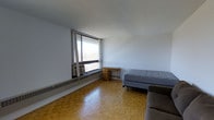 This single-floor space offers a straightforward, functional layout centered around one open living area. The room features classic parquet hardwood flooring in a warm honey tone, adding a timeless character to the interior. A large picture window dominates one wall, allowing generous natural light to fill the space and providing views of the surrounding outdoor landscape. Beneath the window sits a window-mounted air conditioning unit alongside a baseboard heating system, ensuring year-round climate comfort.

The white walls and white ceiling contribute to a clean, minimalist aesthetic, enhancing the sense of openness within the room. The space currently accommodates a bed, a compact wooden desk with a small dresser, and a partial view of an upholstered seating piece, demonstrating its capacity to function as a combined sleeping and living area. The simple, uncluttered design makes the space adaptable to a variety of furniture arrangements and personal needs.

Overall, the space presents a practical and well-lit environment with durable flooring and essential climate control features, suited for straightforward, everyday living.