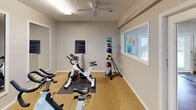 This fitness center is a purpose-built exercise facility designed to accommodate a variety of workout styles and fitness goals. The space is finished with durable rubber tile flooring throughout, providing cushioning and traction for high-impact activities. Several spin bikes are positioned across the main workout area, complemented by a treadmill and an elliptical machine visible through an interior pass-through window that connects to an adjacent cardio room. A vertical medicine ball rack holds an assortment of weighted balls, while stability balls and yoga mats are neatly stored along the walls for flexibility and core training. Large framed mirrors line one wall, creating a sense of openness and allowing users to monitor their form during exercise. The textured ceiling features a ceiling fan that promotes air circulation, and fluorescent lighting ensures the space is well illuminated. A wall-mounted television offers entertainment during workouts. French doors open to an adjoining area, enhancing the flow between spaces. Posted health center guidelines keep users informed of facility rules. The overall atmosphere is clean, functional, and well-organized, making this fitness center a practical and inviting amenity for regular exercise.
