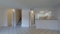 This two-floor residence offers a practical and well-organized layout designed for comfortable everyday living. The main level features large, light-colored ceramic tile flooring that extends throughout the open living area, creating a clean and cohesive look. The space flows naturally toward a kitchen highlighted by white upper cabinetry, granite-style countertops, and a raised breakfast bar that provides both a casual dining option and a natural visual divider between the kitchen and living area. A black refrigerator anchors the kitchen, while recessed and flush-mount ceiling lighting ensures the space remains bright and functional. Adjacent to the living area, a bifold-door closet offers convenient storage access. A carpeted staircase with a handrail leads to the upper floor, adding a warm textural contrast to the tiled lower level. The walls throughout are finished in a neutral white, providing a versatile backdrop that complements a variety of décor styles. Ceiling vents are positioned for efficient climate control across the main level. Overall, the space presents a straightforward, move-in-ready environment with a clean aesthetic and thoughtful layout suited to a range of living needs.