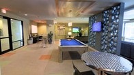 This single-floor amenity space is designed as a multifunctional lounge and recreation area, offering a well-appointed environment for leisure and social gatherings. The centerpiece of the room is a sleek, modern pool table with a bold blue felt surface and a brushed metal frame, positioned beneath a decorative tray ceiling with warm accent lighting that adds architectural interest overhead.

A sculptural stone accent wall flanks one side of the room, providing a dramatic textural contrast to the otherwise smooth, neutral-toned walls. Flat-screen TVs are mounted throughout the space, ensuring visibility from multiple vantage points. High-top tables with metal bar stools are arranged in a casual dining or social area nearby, complemented by computer workstations along the perimeter — suggesting a dual-purpose setup for both entertainment and productivity.

Toward the rear, a comfortable lounge seating area with soft furnishings creates a more relaxed retreat. Recessed lighting and pendant fixtures illuminate the space evenly, while potted plants introduce a touch of greenery. The overall ambiance is modern and inviting, with a cohesive palette of warm greens, neutrals, and metallic finishes throughout.