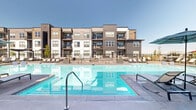 Located at 1595 Twin Lakes Ave in Bozeman, Montana, this outdoor amenity area offers a resort-caliber experience within a contemporary residential community. At its centerpiece is a generously sized swimming pool with crystal-clear blue water, complemented by a separate hot tub/spa nestled nearby. The surrounding deck is finished in clean, light-toned concrete and outfitted with an extensive collection of chaise lounges, shaded by large tilt umbrellas, providing ample space for sunbathing beneath Montana's open skies. A dedicated fire pit lounge area features comfortable outdoor sofas and chairs arranged for gathering, while a covered cabana-style seating nook with lush planters adds a refined touch. Decorative raised planters with ornamental succulents and greenery are integrated throughout the space. The pool area is fully enclosed by sleek black metal fencing, ensuring privacy and safety. Backing the amenity space is a striking multi-story building clad in a mix of board-and-batten siding, horizontal lap siding, and stone accents, with resident balconies overlooking the pool. A separate community clubhouse building with warm wood cladding, stone detailing, and large windows anchors one end of the deck, offering direct access to indoor amenities. The entire space benefits from expansive open sky views characteristic of the Bozeman landscape.