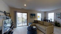 Located at 700 Vista Dr in Gahanna, Ohio, this single-floor residence offers a thoughtfully designed living space with a warm and welcoming ambiance. The living room serves as the heart of the home, bathed in natural light streaming through large windows and a full-width sliding glass door. The sliding door opens directly onto a private balcony, where outdoor seating creates an inviting extension of the living area with pleasant exterior views. Inside, plush wall-to-wall carpeting adds comfort underfoot, while recessed lighting in the ceiling provides clean, even illumination throughout the space. The living room is furnished with a generous sectional sofa and a sleek round coffee table, creating a cozy yet functional gathering area. Neutral wall tones and soft décor accents — including a bold abstract canvas in blue and gold — lend a modern yet approachable aesthetic. A side area near the window accommodates a small dining or accent space, complete with additional décor. The overall layout is open and uncluttered, making the space feel airy and spacious. This residence in Gahanna combines practical single-floor living with an inviting interior atmosphere and easy outdoor access.