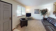 This three-floor space presents a functional and straightforward layout designed for comfortable everyday living. The living area features wall-to-wall carpeting, neutral-toned walls, a textured ceiling, and a large window fitted with horizontal blinds that allows natural light to filter in while providing views of the surrounding greenery. Baseboard heating units are found throughout, providing consistent warmth across the space. The entry area includes a six-panel door flanked by wide windows, an additional wall-mounted AC unit, and durable tile flooring that transitions into the carpeted living space.

The kitchen is equipped with white cabinetry accented by wood-toned framing, ample upper and lower storage, light countertops arranged in a U-shape, a double sink, and an electric range with overhead ventilation. Tile flooring completes the kitchen's clean, utilitarian aesthetic.

Multiple bedrooms feature carpeted floors, baseboard heating, and windows that bring in natural light. One bedroom includes a built-in shelving unit alongside closet space, adding practical storage. The bathrooms are simply appointed, with tile flooring and a tub-shower combination in the full bath, while a secondary bathroom offers a more compact layout with essential fixtures.