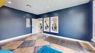 Located at Harper's Crossing in Langhorne, PA, this two-floor fitness facility offers a comprehensive and thoughtfully designed exercise environment. The signature checkered tile flooring—alternating between warm tan and slate gray—runs throughout both levels, lending a clean, classic aesthetic to the space.

The main exercise room is expansive and filled with natural light from large double-hung windows lining multiple walls. It houses a full array of cardio equipment, including treadmills, stair climbers, ellipticals, and stationary bikes, alongside cable weight machines, a dumbbell rack, and a floor-length mirror wall. Structural columns with crown molding detailing add architectural character, while ceiling fans and recessed lighting ensure a comfortable environment year-round.

A second exercise room, accented with bold navy blue walls, serves as a dedicated studio space for stretching, yoga, and functional training. It features exercise mats, medicine balls, and foam rollers displayed on a rack, along with interior windows that overlook the adjacent equipment room—creating a connected yet distinct atmosphere.

An open hallway with sleek black metal railings bridges the two floors, offering elevated views of the cardio floor below. The overall design balances functionality with a polished, community-facility aesthetic suited for active, everyday use.