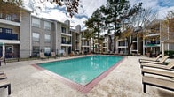 Located at 4500 Sherwood Common Blvd in Baton Rouge, Louisiana, this outdoor amenity area serves as the centerpiece of a well-established residential community. The space is defined by a generously sized swimming pool with clear, blue-green water, framed by a brick-edged concrete deck that provides ample room for relaxation and recreation. Surrounding the pool is a collection of sling-style lounge chairs arranged to take full advantage of the open sky and natural light, making it an ideal spot for outdoor leisure. A shaded seating area with a green market umbrella and patio furniture offers a comfortable retreat for those seeking shelter from the sun. The pool area is enclosed by a black iron fence, providing both security and a clean visual border between the amenity space and the broader grounds. Mature trees — including tall pines and deciduous varieties — provide natural shade and a sense of established greenery throughout the courtyard. Potted plants and manicured shrubs add further landscaping detail along the perimeter. The surrounding multi-story residential buildings feature taupe siding, brick accents, and private balconies that overlook the pool area, creating an enclosed, community-oriented atmosphere. The overall ambiance is inviting and well-maintained, reflecting a comfortable outdoor living environment.
