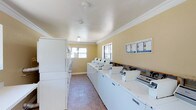 This single-floor communal laundry facility is designed with practicality and efficiency in mind. The room features a generous row of top-load washing machines lined along one wall, complemented by a bank of stacked dryers along the opposite side, providing ample capacity for multiple users simultaneously. The space is finished with warm yellow-toned walls accented by crisp white crown molding, lending a clean and welcoming atmosphere. Light tile flooring runs throughout, making maintenance straightforward and keeping the environment bright. Natural light filters in through multiple windows positioned along the walls, reducing the reliance on artificial lighting and creating an airy, pleasant setting. Overhead lighting ensures the space remains well-lit at all times. A wooden folding or utility counter runs along one wall, offering a convenient surface for sorting and folding laundry. Informational postings are mounted near the machines, providing users with clear instructions and usage guidelines. The overall layout is thoughtfully organized to allow comfortable movement between machines, making this laundry facility a functional and user-friendly amenity.