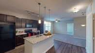 This 1-bedroom, 1-bathroom residence is designed with an open-concept floor plan that seamlessly connects the kitchen, dining, and living areas into a cohesive, functional living space. The kitchen serves as a central hub, anchored by a granite-topped island with an undermount sink and stylish pendant lighting overhead. Dark espresso cabinetry lines the walls, complemented by black appliances including a range, microwave, and refrigerator, as well as track lighting that illuminates the workspace. Wood-style flooring flows through the kitchen and entry areas, transitioning to plush carpet in the living room, where a ceiling fan adds both comfort and visual interest. Natural light pours in through large windows dressed with blinds, brightening the open living area. The bedroom is a quiet, carpeted retreat featuring its own ceiling fan and a generously sized window with views to the exterior. The full bathroom offers a granite vanity countertop, dark cabinetry for storage overhead, a combined tub and shower, and coordinating brushed nickel fixtures. Neutral wall tones throughout create a clean, versatile backdrop that ties the entire space together with a calm, modern ambiance.