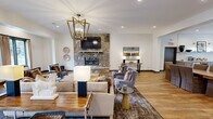 Located at Eagle Pointe Drive in Knoxville, Tennessee, this generously sized single-floor community clubhouse offers a well-appointed amenity space designed for both relaxation and productivity. The main lounge serves as the centerpiece of the facility, showcasing warm hardwood-style flooring, a dramatic floor-to-ceiling stone fireplace, a wall-mounted television, and a gold geometric chandelier that adds a refined touch to the open, airy layout. Natural light filters in through multiple windows, enhancing the inviting atmosphere. An adjacent seating area provides a comfortable business lounge with workstation desks and casual seating, ideal for remote work or quiet reading. The kitchen area is thoughtfully designed with shaker-style cabinetry in a soft gray finish, stone countertops, a classic white subway tile backsplash, brass sconce lighting, a farmhouse sink, and a large island with bar-height seating — perfect for community gatherings. The expansive fitness center is a standout feature, outfitted with treadmills, ellipticals, rowing machines, a full rack system, a punching bag, a comprehensive dumbbell collection, and floor-to-ceiling mirrors. Durable carpet tile flooring and ceiling fans complete the well-ventilated workout space, making this clubhouse a comprehensive community hub.