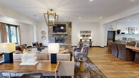 Located at Eagle Pointe Drive in Knoxville, Tennessee, this generously sized single-floor community clubhouse offers a well-appointed amenity space designed for both relaxation and productivity. The main lounge serves as the centerpiece of the facility, showcasing warm hardwood-style flooring, a dramatic floor-to-ceiling stone fireplace, a wall-mounted television, and a gold geometric chandelier that adds a refined touch to the open, airy layout. Natural light filters in through multiple windows, enhancing the inviting atmosphere. An adjacent seating area provides a comfortable business lounge with workstation desks and casual seating, ideal for remote work or quiet reading. The kitchen area is thoughtfully designed with shaker-style cabinetry in a soft gray finish, stone countertops, a classic white subway tile backsplash, brass sconce lighting, a farmhouse sink, and a large island with bar-height seating — perfect for community gatherings. The expansive fitness center is a standout feature, outfitted with treadmills, ellipticals, rowing machines, a full rack system, a punching bag, a comprehensive dumbbell collection, and floor-to-ceiling mirrors. Durable carpet tile flooring and ceiling fans complete the well-ventilated workout space, making this clubhouse a comprehensive community hub.
