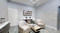 This single-floor space presents a clean, contemporary aesthetic defined by soft grey walls, a suspended drop ceiling with a circular flush-mount light fixture, and a patterned area rug that anchors the seating arrangement. The lounge area is thoughtfully arranged with two cream upholstered swivel chairs facing a low, natural wood coffee table, creating an intimate setting suited for conversation or informal meetings. A white bar-height table with a natural wood base serves as a secondary focal point along the rear wall, accompanied by matching white bar stools and accented by small potted greenery. A framed piece of wall art featuring circular motifs in warm earth tones adds visual interest and a touch of sophistication above the bar table. The white-paneled door provides clean architectural detailing consistent with the overall refined, neutral interior. The space balances functionality and comfort, with its coordinated furnishings, subtle decor, and well-considered layout contributing to a polished, welcoming ambiance well-suited for lounging, entertaining, or professional use.