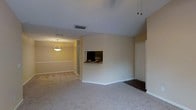 This single-floor residence offers a straightforward and functional layout with interconnected living spaces that flow naturally from one area to the next. The main living area is finished with plush wall-to-wall carpeting in a neutral tone, contributing to a comfortable and understated atmosphere. A textured ceiling runs throughout, and HVAC vents are conveniently positioned overhead for efficient climate control. A key architectural feature is the built-in pass-through opening in the dividing wall, which connects the living area to an adjacent space — ideal for maintaining visual connectivity between rooms. A decorative pendant light hangs in the adjoining room, adding a warm, focused glow that complements the soft, neutral wall tones found throughout. Toward the right side of the space, a transition to dark hardwood-style flooring marks the entry area near what appears to be a front or utility door, offering a practical and durable surface at the threshold. White baseboards frame each room cleanly, and the overall color palette of warm beige and off-white walls creates a bright, welcoming environment. The open sightlines and connected layout make the space feel expansive and easy to navigate.