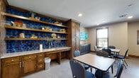 This single-floor community space is thoughtfully designed to balance rustic charm with functional amenities, making it well-suited for group gatherings, activities, and socializing. The entrance features rich, dark wood-framed doors alongside glass double doors that open into the main area, creating an inviting and warm transition into the space.

The kitchenette area serves as a standout focal point, showcasing a striking deep-blue glazed tile backsplash that spans the full width of the wall. Rich wood cabinetry with ample storage, open display shelving, and a granite-topped counter with a built-in sink complete this functional yet visually compelling area. A wall-mounted television and additional storage cabinetry are positioned nearby for added convenience.

The main gathering room is generously sized to accommodate multiple seating arrangements with tables and chairs positioned throughout. A distinctive textured stone-look accent wall adds an organic, earthy character to the room, while a second wall-mounted television enhances its versatility. Rustic wood-framed doors with decorative ironwork provide access to additional areas, contributing to the cohesive design language found throughout.

Natural light filters in through multiple windows, brightening the light-toned flooring and neutral walls. Recessed lighting ensures the space remains well-lit at all times. The overall ambiance is warm and welcoming, combining rustic architectural details with modern, practical features ideal for community use.