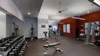 The fitness center is a thoughtfully designed workout space that combines functionality with a dynamic aesthetic. The room features a striking color scheme, with a bold burnt-orange accent wall providing visual contrast against the neutral gray walls and dark rubberized flooring, creating an energetic atmosphere conducive to exercise.

The space is well-lit through a combination of recessed ceiling lights and suspended linear fluorescent fixtures, ensuring even illumination throughout. A ceiling fan contributes to air circulation and comfort during workouts. Natural light filters in through slatted blinds on the windows along the accent wall, adding brightness to the far end of the room.

Equipment is organized to maximize the available floor space. A rack of dumbbells lines one wall, offering a full range of free weights for strength training. Multiple adjustable workout benches are positioned centrally for versatile use. Cardio machines, including stationary bikes and other equipment, are arranged toward the rear of the space. A cable machine provides additional strength-training options, while medicine balls on a dedicated rack round out the amenities. A mounted television is also available for entertainment during workouts.