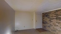 This single-floor space presents a straightforward, functional layout with several notable design elements. The interior is finished with smooth, neutral-toned walls that create a clean and understated backdrop throughout the main living area. A standout architectural feature is the exposed brick accent wall positioned near the entryway, adding texture and a rustic character that contrasts with the otherwise modern, minimalist interior.

The flooring transitions from carpet in the main living area to what appears to be hard flooring near the entry, providing a practical delineation between zones. The drop-tile ceiling gives the space a classic, traditional aesthetic commonly found in residential and light commercial properties. A standard white interior door serves as the main entry point, flanked by the brick wall on one side.

Electrical outlets and a wall switch plate are visible along the walls, indicating accessible utility placements. The space is currently unfurnished, allowing for a clear view of its proportions and layout. Overall, the interior offers a blank canvas with a hint of character through its exposed brick detail, suitable for a variety of uses and personalization.