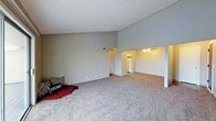 This single-floor space is defined by its striking vaulted ceilings, which rise steeply to create a sense of expansive vertical volume in the main living area. The soaring ceiling height adds an airy, open quality that makes the space feel notably larger than its footprint suggests. Neutral gray walls complement the space throughout, providing a clean, understated backdrop.

The main living area is finished with wall-to-wall carpet in a warm beige-gray tone, contributing to a comfortable and casual atmosphere. The open layout allows for flexible furniture arrangement, with ample room to define separate functional zones within the space.

Transitioning from the living area, a lower-ceilinged section is visible toward the rear, housing what appears to be a kitchen. This area is outfitted with a ceiling fan and light fixture, and a white refrigerator is visible, suggesting a functional kitchen layout. The transition between the vaulted living space and the adjacent area is framed by a half-wall partition, adding subtle architectural definition.

Overall, the space presents a practical and open design, with the dramatic ceiling height serving as its most notable architectural feature.