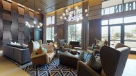 This contemporary lobby makes an immediate impression with its soaring double-height ceilings, floor-to-ceiling windows, and an abundance of natural light that floods the main entrance. The space is thoughtfully divided into several distinct zones, each with its own character and purpose. At the heart of the lobby, a stylish lounge area is anchored by a bold geometric blue-and-white area rug, surrounded by an eclectic mix of wingback chairs, upholstered armchairs, and a leather ottoman, all illuminated by striking globe chandelier fixtures. Warm hardwood flooring flows throughout, contrasting beautifully with large-format light tile in the transitional corridors. A secondary seating area features a built-in linear fireplace framed by dark stone cladding and integrated shelving, complemented by a plush sectional sofa and accent chairs. Deep blue textured walls serve as a dramatic backdrop in multiple areas, accented by curated, illuminated artwork in recessed niches. Sculptural three-dimensional wall panels add architectural depth and visual intrigue. Near the elevator vestibule, a comfortable banquette lounge with a padded ribbed wall panel and colorful ottoman seating offers a more intimate waiting area. Rich walnut wood paneling, recessed cove lighting, and a carefully layered material palette of metal, stone, and wood unify the entire space into a cohesive, upscale aesthetic.