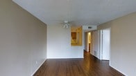 This single-floor residence offers a clean, functional layout designed for comfortable everyday living. The main living area is an open, well-proportioned space finished with rich, dark hardwood-style flooring that runs throughout, adding warmth and a sense of continuity. A ceiling fan with integrated lighting is centrally positioned, providing both air circulation and ambient illumination beneath a textured ceiling. Neutral-toned walls keep the space feeling bright and versatile, ready to accommodate a variety of interior styles. A distinctive pass-through window connects the living area directly to the kitchen, which features warm wood cabinetry and a bright overhead light, creating an open, communicative flow between the two spaces. This architectural detail makes the kitchen feel accessible while maintaining a defined separation of function. A hallway extending from the living area leads to additional rooms, with closet storage clearly visible — a practical feature that supports organized living. Flush-mounted HVAC vents are integrated into both the floor and ceiling, ensuring efficient climate control throughout. The overall ambiance is understated and practical, with a layout that emphasizes ease of movement and livability.
