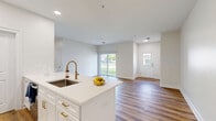 Located at 60 Weibel Ave in Saratoga Springs, NY, this well-appointed 2-bedroom, 2-bathroom single-floor home offers a thoughtfully designed layout with modern finishes throughout. The open-concept living room and kitchen area serves as the heart of the home, featuring warm hardwood-style flooring, recessed lighting, and a sliding glass door that floods the space with natural light and provides outdoor views. The kitchen is a standout, boasting crisp white cabinetry, stainless steel appliances including a refrigerator, range, and over-the-range microwave, and a large center island with a quartz countertop and undermount sink — ideal for both cooking and casual dining. Both bedrooms are generously proportioned, featuring the same hardwood-style flooring and large windows that invite ample natural light. The primary bedroom includes a double-door closet for convenient storage. The two bathrooms are finished with light tile flooring, white vanities with gold-toned hardware, and clean, bright aesthetics. A utility room adds practical functionality, while the attached garage provides secure parking and additional storage space. The home's neutral color palette and modern design elements create a cohesive, move-in-ready atmosphere throughout.