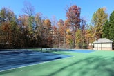 This single-level outdoor space is defined by its well-maintained tennis courts and inviting covered porch area, set against a backdrop of mature trees displaying rich autumn colors. The tennis courts feature a classic green and blue hard surface finish, complete with regulation nets, clearly marked court lines, and multiple sets of overhead lighting towers positioned throughout, allowing for evening play. A perimeter fence encloses the courts, providing both safety and boundary definition, while a small outbuilding is visible at the court's edge, likely serving as storage or a utility structure.

Adjacent to the courts, a spacious covered porch offers a refined gathering area characterized by elegant white classical columns rising from substantial natural stone base piers. A low stone perimeter wall frames the porch, complementing the overall craftsmanship and adding a sense of permanence to the structure. The concrete flooring underfoot is clean and well-kept. Mature landscaping, including neatly trimmed shrubs and natural ground cover, surrounds the porch area, seamlessly blending the built environment with the wooded setting. The combination of the courts and covered porch creates a versatile recreational and social environment.
