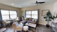 This well-appointed single-floor residence offers a thoughtfully designed living space that blends comfort and contemporary style. The living area serves as the heart of the home, showcasing rich hardwood-style flooring that anchors the room with warmth and elegance. Large windows dressed with soft blue curtains flood the space with natural light, creating an open and airy atmosphere throughout. A neutral color palette of warm beiges and grays provides a calming backdrop that complements the carefully curated furnishings. The living room features a plush sectional sofa in a muted gray tone, paired with a geometric-patterned area rug and a sleek coffee table, creating an inviting and functional seating arrangement. Accent chairs and a decorative pouf add layers of texture and visual interest. Recessed lighting and a modern ceiling fan with a dark wood finish enhance both the functionality and aesthetic of the space. Tasteful wall art and decorative accents, including a sideboard and potted greenery, lend a polished, curated feel to the interior. The overall ambiance is relaxed yet refined, making this space ideal for both everyday living and entertaining guests.