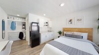 Located at 311 W Trinity Lane in Nashville, Tennessee, this single-floor studio unit offers a clean, modern design with a bright, open layout. The main living area seamlessly integrates the sleeping and kitchen spaces, finished with warm wood-look luxury vinyl plank flooring and crisp white walls that create a light and airy atmosphere throughout.

The kitchenette is thoughtfully appointed with white shaker-style cabinetry, open shelving, a light speckled stone countertop, a stainless steel undermount sink, and a full-size black refrigerator with a microwave stacked on top — providing all the essential amenities for everyday living. A wall-mounted flat-screen TV and a compact fold-down desk with a chair offer practical spaces for entertainment and productivity.

The sleeping area features a natural wood-paneled headboard that adds warmth and texture to the neutral palette, complemented by recessed lighting overhead. A windowed entry door allows natural light to flow into the space.

The private bathroom is finished in a consistent, clean style, featuring a tiled walk-in shower with a curtain enclosure, a white vanity with countertop, and wood-look flooring that ties back to the main living area. A walk-in closet with shelving and hanging space provides ample storage, completing this efficiently designed and well-appointed studio.