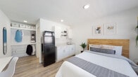 Located at 311 W Trinity Lane in Nashville, Tennessee, this single-floor studio unit offers a clean, modern design with a bright, open layout. The main living area seamlessly integrates the sleeping and kitchen spaces, finished with warm wood-look luxury vinyl plank flooring and crisp white walls that create a light and airy atmosphere throughout.

The kitchenette is thoughtfully appointed with white shaker-style cabinetry, open shelving, a light speckled stone countertop, a stainless steel undermount sink, and a full-size black refrigerator with a microwave stacked on top — providing all the essential amenities for everyday living. A wall-mounted flat-screen TV and a compact fold-down desk with a chair offer practical spaces for entertainment and productivity.

The sleeping area features a natural wood-paneled headboard that adds warmth and texture to the neutral palette, complemented by recessed lighting overhead. A windowed entry door allows natural light to flow into the space.

The private bathroom is finished in a consistent, clean style, featuring a tiled walk-in shower with a curtain enclosure, a white vanity with countertop, and wood-look flooring that ties back to the main living area. A walk-in closet with shelving and hanging space provides ample storage, completing this efficiently designed and well-appointed studio.