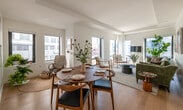 This contemporary single-floor residence is designed with a clean, modern aesthetic throughout. Light hardwood flooring and crisp white walls create a bright, cohesive atmosphere across every room. The open-plan living and dining area is a standout feature, with oversized black-framed windows on multiple sides flooding the space with natural light and offering expansive urban skyline views. The living area comfortably accommodates a full seating arrangement and flows seamlessly into a well-defined dining space, making it ideal for both relaxed living and entertaining.

The kitchen is thoughtfully designed with dark walnut lower cabinetry, sleek matte upper cabinets, white countertops, and under-cabinet lighting that adds a refined touch. It includes built-in appliances such as an oven, induction cooktop, and microwave, all set within an efficient U-shaped layout.

The primary bedroom is generously sized, featuring large windows with city views and ample wall space. A secondary bedroom, accessible via a glass-paneled door, offers its own windows with natural light. The bathroom includes a full soaking tub with a tiled surround in a classic white subway pattern. Throughout the residence, tray ceilings add architectural dimension, contributing to the overall sense of height and sophistication.