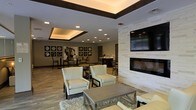 This well-appointed single-floor space serves as a sophisticated communal gathering area, thoughtfully designed to balance comfort with modern elegance. The lounge area anchors the room with a striking floor-to-ceiling stacked stone fireplace, topped by a large wall-mounted television — creating a natural focal point for relaxation and socializing. Plush cream armchairs and a sleek metallic coffee table are arranged atop a soft area rug, offering an inviting seating arrangement on warm hardwood-style flooring.

Overhead, a coffered tray ceiling with integrated cove lighting and recessed fixtures casts a warm, layered glow throughout the space, adding architectural depth and a sense of refined detail. Dark wood trim accents frame the ceiling, complementing the rich tones found throughout the room.

Toward the back, a dining area features multiple tables and chairs arranged beneath decorative wall art — a curated grid of circular framed pieces that adds a gallery-like quality to the space. A counter or bar area is visible in the background, suggesting additional amenities for guests or residents. The overall ambiance is warm, modern, and welcoming, making this space well-suited for both casual gatherings and more structured social events.