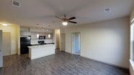 This single-floor residence features an open-concept layout that connects the kitchen and living area into one cohesive, functional space. The kitchen is outfitted with modern taupe cabinetry, black appliances including a built-in microwave, and a central island with a sink that serves as both a prep area and a natural divider between the kitchen and living space. Stylish pendant lights hang above the island, complementing the recessed and track lighting installed throughout the kitchen for practical illumination. The living area is spacious and unobstructed, anchored by a ceiling fan that promotes airflow and adds a casual, comfortable touch to the room. Durable wood-style plank flooring runs continuously throughout, lending a clean and contemporary look. The neutral, light-toned walls keep the space feeling bright and open. Two doors visible from the main living area lead to additional rooms, suggesting a thoughtful floor plan with private spaces set apart from the main living zone. Overall, the layout balances an airy, open communal area with clearly defined private spaces.
