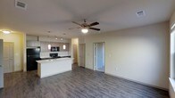 This single-floor residence features an open-concept layout that connects the kitchen and living area into one cohesive, functional space. The kitchen is outfitted with modern taupe cabinetry, black appliances including a built-in microwave, and a central island with a sink that serves as both a prep area and a natural divider between the kitchen and living space. Stylish pendant lights hang above the island, complementing the recessed and track lighting installed throughout the kitchen for practical illumination. The living area is spacious and unobstructed, anchored by a ceiling fan that promotes airflow and adds a casual, comfortable touch to the room. Durable wood-style plank flooring runs continuously throughout, lending a clean and contemporary look. The neutral, light-toned walls keep the space feeling bright and open. Two doors visible from the main living area lead to additional rooms, suggesting a thoughtful floor plan with private spaces set apart from the main living zone. Overall, the layout balances an airy, open communal area with clearly defined private spaces.