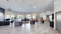 This well-appointed community clubhouse offers a generous, open-plan layout spread across a single primary floor. The main lounge area serves as the heart of the space, featuring light hardwood-style flooring, recessed ceiling lighting, and an abundance of natural light streaming through large windows that frame views of the outdoor pool. The lounge is furnished with plush, tufted sofas and a central coffee table, creating a comfortable social gathering zone. A dedicated dining and café area sits adjacent, complete with bold orange accent chairs, sleek dark tables, and a beverage station set against a white subway tile backdrop. A wall-mounted flat-screen TV and arched entryways add architectural character throughout. The entrance hallway makes a strong first impression with a dramatic dark fluted-panel accent wall, a reception desk, and elegant decorative touches. The exercise room is fully equipped with treadmills, ellipticals, a free-weight rack, and cardio machines, all complemented by high vaulted ceilings, large windows, and a striking motivational mural. Outside, a resort-style pool surrounded by lounge chairs, palm trees, and a pergola-shaded seating area completes this vibrant amenity space.