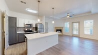 This thoughtfully designed single-floor residence in Lenexa, Kansas offers a seamless open-concept layout that connects the kitchen and living areas into one cohesive, light-filled space. The kitchen is a standout feature, showcasing a large center island with a white quartz countertop and undermount sink, two-tone cabinetry with white uppers and charcoal lower cabinets, a classic subway tile backsplash, and a full suite of stainless steel appliances. Elegant pendant lighting adds a refined touch above the island. The living room impresses with a tray ceiling, ceiling fan, a tile-surround fireplace, and access to the outdoors through multiple windows that invite generous natural light. Light-toned wood-style flooring flows consistently throughout the main living areas, enhancing the bright and airy ambiance. A dedicated laundry area includes an in-unit front-loading washer. The bathroom is well-appointed with a soaking tub and shower combination, a vanity with ample storage, and a connected closet with wire shelving. Additional louvered closet doors provide practical storage solutions. The overall design reflects a clean, contemporary aesthetic with attention to functional detail.