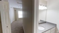 This single-floor unit presents a straightforward, functional layout with distinct living and bathroom areas connected by a central corridor. The main living area is finished with wall-to-wall carpet and features white walls that contribute to a clean, neutral aesthetic. Natural light enters through a window, brightening the open space, which also showcases exposed overhead piping and industrial-style ceiling elements that lend the unit a subtle loft-like character. The ceiling height adds to the sense of openness throughout this area.

Adjacent to the living space, the bathroom is equipped with a well-lit vanity featuring a built-in sink and a wide mirror framed by a row of globe lights overhead, providing ample illumination. The countertop offers functional surface space, and the overall finish is clean and utilitarian. A separate area visible in the floor plan suggests additional functional zones, including what appears to be a kitchen or utility space. The unit's layout is compact yet efficiently organized, making practical use of the available footprint. The consistent use of white and neutral tones throughout creates a cohesive and understated ambiance suited for a variety of uses.