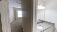 This single-floor unit presents a straightforward, functional layout with distinct living and bathroom areas connected by a central corridor. The main living area is finished with wall-to-wall carpet and features white walls that contribute to a clean, neutral aesthetic. Natural light enters through a window, brightening the open space, which also showcases exposed overhead piping and industrial-style ceiling elements that lend the unit a subtle loft-like character. The ceiling height adds to the sense of openness throughout this area.

Adjacent to the living space, the bathroom is equipped with a well-lit vanity featuring a built-in sink and a wide mirror framed by a row of globe lights overhead, providing ample illumination. The countertop offers functional surface space, and the overall finish is clean and utilitarian. A separate area visible in the floor plan suggests additional functional zones, including what appears to be a kitchen or utility space. The unit's layout is compact yet efficiently organized, making practical use of the available footprint. The consistent use of white and neutral tones throughout creates a cohesive and understated ambiance suited for a variety of uses.