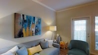 This single-floor space presents a thoughtfully designed living area that balances comfort and style. The living room serves as the heart of the space, anchored by a large, bold abstract canvas in shades of blue, gold, and white that commands attention on the neutral-toned wall. Crown molding along the ceiling adds a refined architectural detail, lending the room a polished, finished look.

The seating area is anchored by a light gray sofa adorned with yellow and white accent pillows, contributing a warm, inviting atmosphere. A teal armchair with floral accent pillows adds a pop of color and visual interest. A tripod floor lamp provides warm ambient lighting, complementing the soft, neutral wall tones throughout.

A side table between the seating pieces offers functional surface space, styled with decorative objects. Natural light filters in through a window fitted with blinds, brightening the space without overwhelming it. The wood-toned flooring grounds the room and adds warmth. Overall, the space conveys a comfortable, modern aesthetic with carefully chosen furnishings and décor that create a cohesive and welcoming environment.
