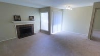 This single-floor residence offers a comfortable and functional layout designed for everyday living. The main living area is a generous, open room finished with wall-to-wall carpeting in a neutral tone, creating a warm and inviting atmosphere. A standout feature of this space is the wood-burning fireplace, framed by a classic tile surround and a clean white mantel — an ideal focal point for the room. Natural light flows generously into the space through a large horizontal window fitted with blinds, as well as a full-length sliding glass door dressed with vertical blinds, which likely leads to an outdoor area and further brightens the interior. The walls are painted in a soft, neutral tone that complements the overall aesthetic and enhances the sense of openness. Overhead, a textured ceiling adds subtle character, while a modern multi-bulb track light fixture provides practical and stylish illumination. White baseboards trim the perimeter of the room, adding a crisp, finished look. Overall, the space presents a clean, move-in ready environment with classic residential features that balance comfort with everyday practicality.