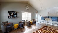 Located at 1700 Woodbury Rd in Orlando, FL, this business center is a well-appointed, single-floor communal workspace designed to accommodate a variety of productivity needs. The space is immediately distinguished by its soaring vaulted ceilings with a clean white tongue-and-groove finish, which lend an airy, open feel throughout.

The interior features rich hardwood-style flooring anchored by a bold, patterned area rug in deep browns and yellows, adding a vibrant design element to the space. Flexible seating is a hallmark of the layout, with numerous mobile chairs in gray and purple tones paired with compact yellow ottoman accents and individual tablet-arm surfaces — ideal for solo work or small group collaboration.

Along one wall, a row of counter-height workstations with blue task chairs provides a dedicated zone for computer use, complemented by decorative wall art above. Opposite, a textured accent wall framed by a rustic wood beam adds warmth and architectural character to the neutral gray palette.

Natural light pours in through large multi-pane windows, brightening the workspace and offering views of the outdoor pool area and surrounding greenery. Double glass doors provide convenient access to the exterior. The overall ambiance is modern, functional, and welcoming — a versatile space well-suited for work, study, or meetings.