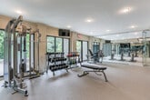 Located at 3000 Cumberland Club Dr in Atlanta, Georgia, this fitness center offers a well-appointed, single-floor workout facility designed to support a comprehensive range of exercise routines. The space is organized into distinct functional zones, beginning with a strength training area that houses a full rack of dumbbells, a colorful set of kettlebells, an adjustable weight bench, and a cable/multi-function resistance machine. Floor-to-ceiling mirrors line the walls, visually expanding the space and allowing exercisers to monitor their form throughout their workouts. A dedicated cycling studio features multiple stationary bikes arranged in front of a wall-mounted flat-screen television, providing an immersive cardio experience. The facility is entered through sleek glass doors with stainless steel hardware, lending a polished, modern aesthetic to the space. Neutral-toned carpeted flooring runs throughout, and recessed lighting ensures a bright, well-lit environment. Large windows and sliding glass doors along the perimeter flood the space with natural light and offer views of lush, green tree canopy, creating a refreshing and energizing atmosphere. A private outdoor terrace is accessible directly from the main workout area, extending the fitness experience to the outdoors.