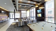 Located at 6963 E 109th Ave in Westminster, Colorado, this single-floor community clubhouse offers a contemporary and inviting gathering space designed for both relaxation and entertainment. The main lounge area is anchored by a full-sized billiards table and complemented by a seating arrangement of striped upholstered armchairs and modern accent furniture, creating a comfortable social atmosphere. Exposed dark wood ceiling beams contrast elegantly against the light ceiling, adding warmth and architectural character to the space. Recessed lighting and stylish pendant fixtures provide layered illumination throughout. A feature wall outfitted with multiple flat-screen televisions serves as a dynamic entertainment focal point, finished in rich dark paneling for a sophisticated look. Floor-to-ceiling windows and what appears to be a large garage-style roll-up window wall flood the interior with natural light while offering views to an outdoor area, seamlessly blending interior and exterior spaces. The open layout accommodates multiple activity zones, from quiet seating clusters to active recreation areas. Patterned carpeting and thoughtful furnishings throughout contribute to a polished, modern aesthetic that makes this clubhouse a standout amenity.