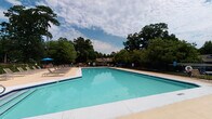 Located at 1025 Avent Hill in Raleigh, NC, this single-story pool clubhouse serves as a central amenity hub for the community. The standout feature is the expansive outdoor swimming pool, its clear turquoise water framed by a generous concrete deck and surrounded by mature trees that provide a natural, shaded backdrop. A row of lounge chairs and umbrella-shaded seating along the poolside offers ample space for relaxation, while a covered pergola structure provides additional shade at the pool's edge. The interior of the clubhouse is clean and functional, featuring warm wood-tone flooring throughout and neutral gray walls accented with nautical-themed artwork. A central hallway connects key amenities, including clearly marked restroom facilities — with both gender-specific and accessible options — and a wall-mounted water fountain for convenience. A small lounge nook off the main corridor provides a comfortable seating area, furnished with a cushioned bench and a potted plant, creating a welcoming spot to rest. Large windows and glass doors connect the interior to the pool area, maintaining a seamless indoor-outdoor flow and filling the space with natural light. Overall, the clubhouse offers a practical, well-appointed environment designed to support an active outdoor lifestyle.