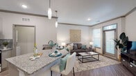 This single-floor residence offers a thoughtfully designed open-concept layout that seamlessly connects the kitchen, dining, and living areas. The kitchen is outfitted with stainless steel appliances, including a range, microwave, and refrigerator, complemented by a subway tile backsplash, granite countertops, and a mix of white upper and gray lower cabinetry. A peninsula with seating extends the countertop space and serves as a casual dining area, illuminated by sleek pendant lights overhead.

The living room is bright and welcoming, featuring wood-style flooring, crown molding, and multiple windows that allow natural light to fill the space. A door provides direct access to the exterior, enhancing the open feel of the room.

The bedroom is carpeted for comfort and includes a ceiling fan, ample natural light from a large window, and sufficient space for a full furniture arrangement. The bathroom features wood-look flooring, a granite-topped vanity with shaker-style cabinetry, a large mirror, and a tub-shower combination with floor-to-ceiling marble-style tile surround. The overall color palette is neutral and cohesive, creating a clean, modern ambiance throughout.