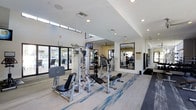 This two-story fitness facility is designed to deliver a comprehensive workout experience in a bright, modern environment. The main gym area is outfitted with a variety of strength training machines, including cable systems, a leg press, and adjustable benches, alongside cardio equipment to support a full range of exercise routines. The space is finished with light-toned flooring accented by blue patterned carpet, contributing to a clean and energetic aesthetic. Large floor-to-ceiling windows line one side of the room, flooding the interior with natural light and offering views of an outdoor pool area, seamlessly blending indoor and outdoor environments. Elevated clerestory windows near the upper portion of the walls further enhance the sense of openness and airiness throughout the space. Full-length mirrors are strategically positioned along the walls to maximize the perception of space and assist exercisers in monitoring their form. Recessed lighting supplements the abundant natural light, ensuring the facility remains well-lit at all times. The high ceilings create a spacious, unencumbered atmosphere ideal for focused training sessions. The overall design strikes a balance between functionality and contemporary style, making this fitness center a well-appointed destination for health and wellness.