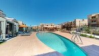 This expansive outdoor amenity space centers around a generously sized freeform pool with crystal-clear turquoise water, designed to evoke a resort-style atmosphere. The pool's curved, organic shape is complemented by a broad stamped concrete deck that surrounds it on all sides, providing ample room for lounging and recreation. A row of private cabanas with flowing curtains and elegant canopies lines one side of the deck, each outfitted with comfortable seating for shade and relaxation. Vibrant orange market umbrellas are strategically positioned throughout the area, adding a pop of color against the warm desert palette of the surrounding multi-story residential buildings. The pool deck is further enhanced by an array of cushioned chaise lounges, ideal for sunbathing in the abundant natural light. A covered patio structure at one end of the pool provides additional sheltered seating. The entire area is enclosed by a sleek wrought iron fence, ensuring both security and unobstructed sightlines. Lush desert landscaping borders the perimeter, integrating the space naturally into its arid environment. The backdrop features dramatic mountain views under brilliantly clear blue skies, reinforcing the open, sun-drenched ambiance that defines this outdoor retreat.