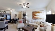 This single-floor residence showcases an open-concept floor plan that connects the living and dining areas in a cohesive, functional layout. The living area is centered around a neutral color palette, with light-toned walls and flooring that create a bright, welcoming atmosphere. A ceiling fan with an integrated light fixture serves as a practical focal point overhead, enhancing air circulation and ambient lighting throughout the space.

The living room accommodates a comfortable seating arrangement, including a plush sofa, patterned accent chair, and an upholstered ottoman that doubles as a coffee table. A bold, abstract canvas with warm rust and ochre tones adds a striking artistic element to the main wall, contributing visual depth to the room.

Transitioning naturally from the living area, the dining space features a dark-toned table paired with matching chairs, ideal for everyday meals or entertaining. A decorative mirror and accent dresser positioned along the wall add character and storage functionality to the transitional space. Throughout, the consistent use of neutral flooring and soft lighting ties the open layout together, creating a harmonious and inviting environment.
